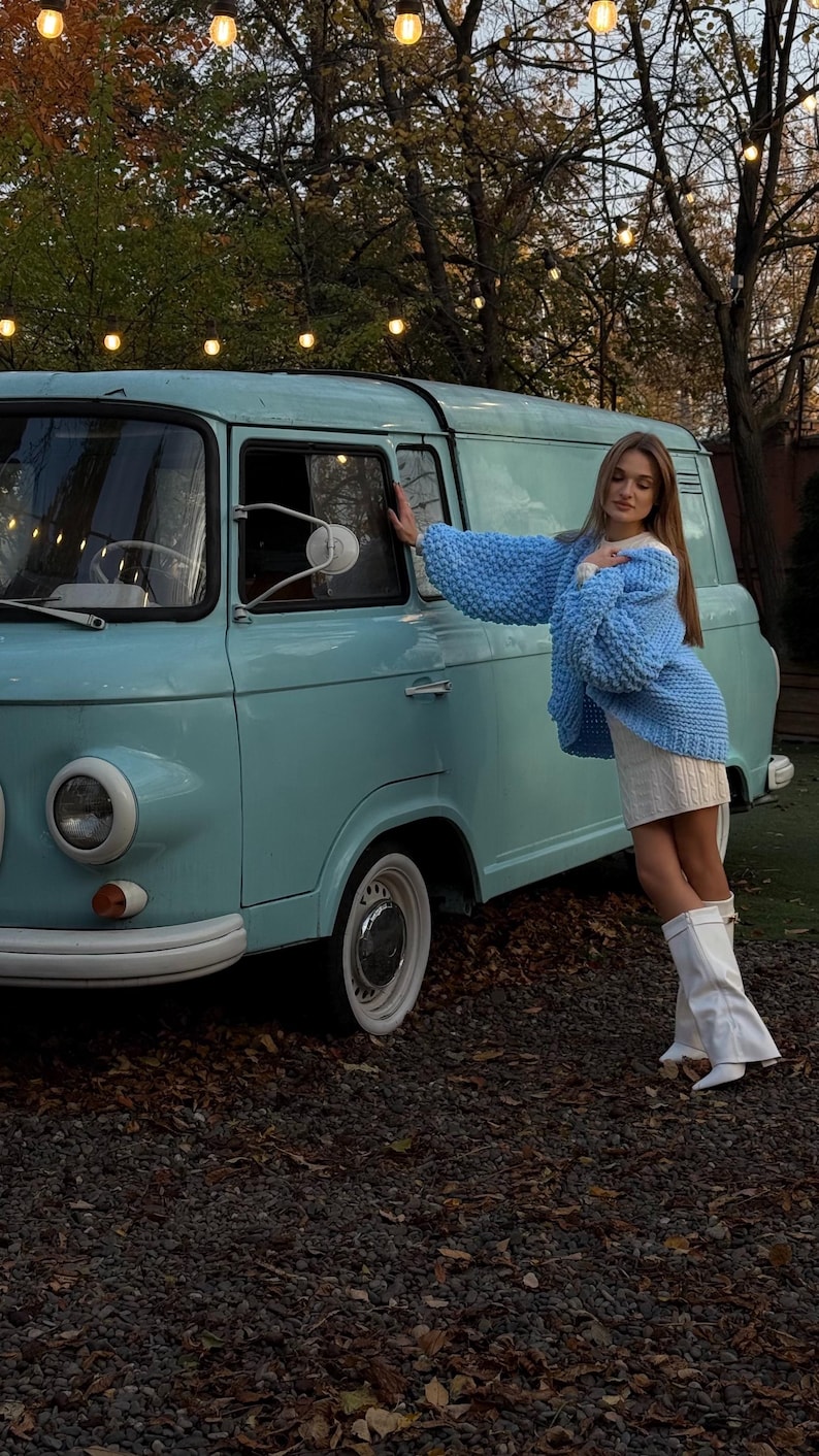 May include: A light blue vintage van with white tires and a white bumper. The van is parked on a gravel surface. A person is wearing a light blue knit sweater and white boots, leaning against the van.