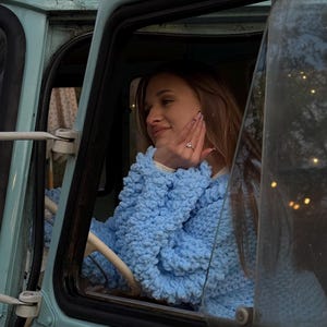 May include: A light blue, vintage-style vehicle with a person wearing a light blue, textured sweater. The person is inside the vehicle, resting their hand on their face. The vehicle's interior is visible through the window.