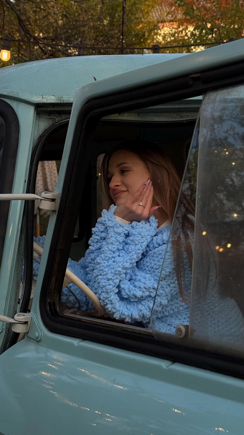 May include: A light blue, vintage-style vehicle with a person wearing a light blue, textured sweater. The person is inside the vehicle, resting their hand on their face. The vehicle's interior is visible through the window.