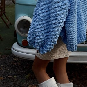 May include: A light blue, chunky knit sweater with oversized sleeves. The sweater is paired with a cream-colored cable knit skirt and white knee-high boots. A light blue vintage van is in the background.