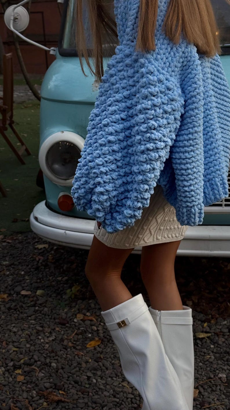 May include: A light blue, chunky knit sweater with oversized sleeves. The sweater is paired with a cream-colored cable knit skirt and white knee-high boots. A light blue vintage van is in the background.