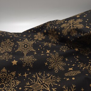 Può includere: Tessuto nero con un motivo ripetuto di fiocchi di neve, stelle e punti dorati. Il tessuto è drappeggiato, creando pieghe e ombre che evidenziano il design. Il tema generale suggerisce l'inverno o le festività.