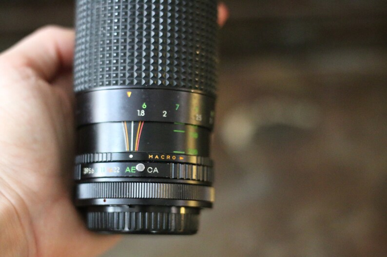 Może przedstawiać: Czarny obiektyw do aparatu z srebrnym pierścieniem i oznaczeniami ustawień przysłony, w tym 1,8, 2, 6 i 7. Obiektyw ma r&oacute;wnież ustawienie makro.