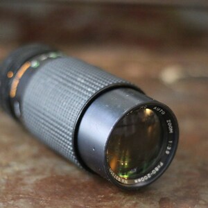 Może przedstawiać: Czarny obiektyw do aparatu z srebrnym pierścieniem i nadrukiem "RBO-2000R ZOOM 1:3.9 AUTO" na boku.