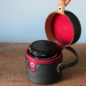 May include: A black camera lens with a UV filter in a black leather case with a red lining and a brown leather strap.