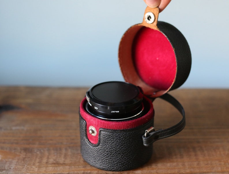May include: A black camera lens with a UV filter in a black leather case with a red lining and a brown leather strap.