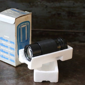 Może przedstawiać: Czarny obiektyw Super Albinar-ADG 35-105mm f3.5 w oryginalnym białym opakowaniu styropianowym. Obiektyw znajduje się w białym pudełku z niebieskim i czarnym tekstem, kt&oacute;ry brzmi "Super Albinar-ADG Multi-Coated Zoom 35-105mm f3.5".