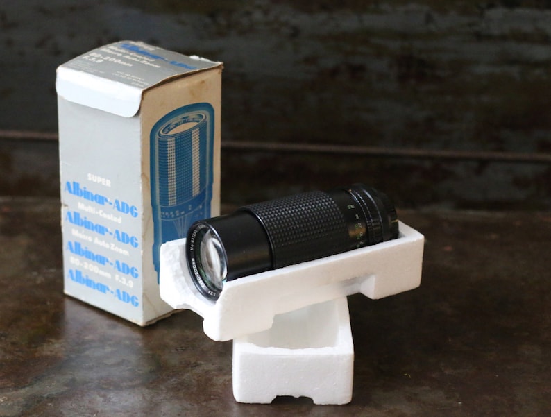 Może przedstawiać: Czarny obiektyw Super Albinar-ADG 35-105mm f3.5 w oryginalnym białym opakowaniu styropianowym. Obiektyw znajduje się w białym pudełku z niebieskim i czarnym tekstem, kt&oacute;ry brzmi "Super Albinar-ADG Multi-Coated Zoom 35-105mm f3.5".