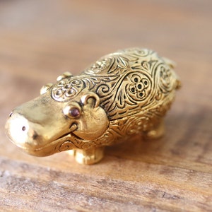 以下が含まれることがあります： 金色の装飾的なカバの置物。複雑な花柄と渦巻き模様が施されています。カバは小さく丸い紫色の目をしています。この置物は金属製で、おそらく収集品です。