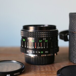 May include: A black camera lens with a silver ring and green markings. The lens has a depth of field scale with numbers from 28 to 22. The lens is in a black leather case.