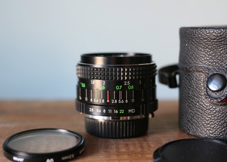 May include: A black camera lens with a silver ring and green markings. The lens has a depth of field scale with numbers from 28 to 22. The lens is in a black leather case.