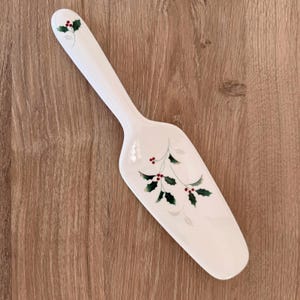 Può includere: Servitore per torte in ceramica bianca con design a bacche di agrifoglio. Il servitore presenta un lungo manico e una lama larga e piatta decorata con foglie verdi e bacche rosse, perfetto per le feste.