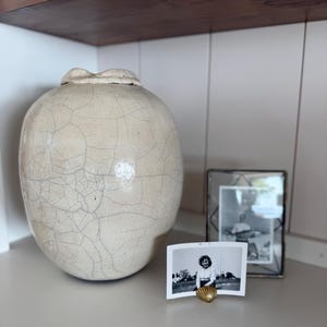 May include: A large, round, off-white ceramic vase with a prominent dark crackle glaze finish. Next to it sits a small, gold-colored seashell-shaped photo holder displaying a black and white photograph, alongside a rectangular metal picture frame.