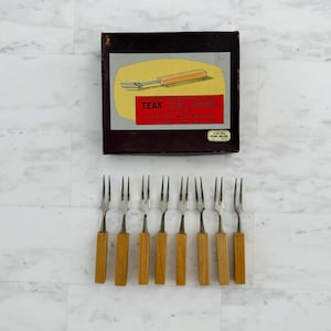 May include: Set of eight teak handled mini forks with stainless steel tines. The forks are arranged in a row on a white marble surface. The box the forks came in is in the background. The box is brown with a yellow label that reads "Teak Tiny Forks" and "Viking Hand Made".