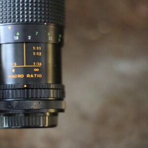Może przedstawiać: Zbliżenie na czarny obiektyw aparatu z skalą makro. Skala zawiera oznaczenia dla 1:5, 1:11, 1:12, 1:16 i nieskończoności.