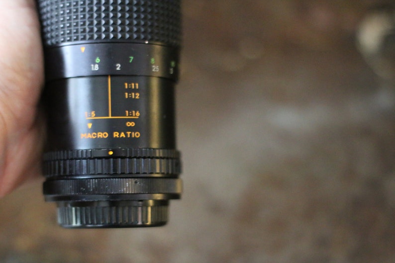 Może przedstawiać: Zbliżenie na czarny obiektyw aparatu z skalą makro. Skala zawiera oznaczenia dla 1:5, 1:11, 1:12, 1:16 i nieskończoności.