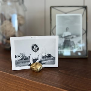May include: A black and white photograph of a woman in a white blouse and dark skirt, propped up by a gold seashell. Another framed photo and a glass jar with decorative objects are in the background. The photo is dated May '61.
