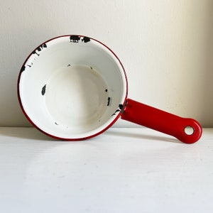 以下が含まれることがあります： 赤い取っ手がついた白いエナメル製の鍋。鍋は、白いエナメルに黒い斑点があり、使い古されたビンテージな外観をしています。