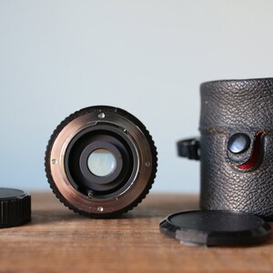 May include: A black camera lens with a silver ring around the front. The lens is sitting on a wooden surface with a black leather case and a black lens cap.
