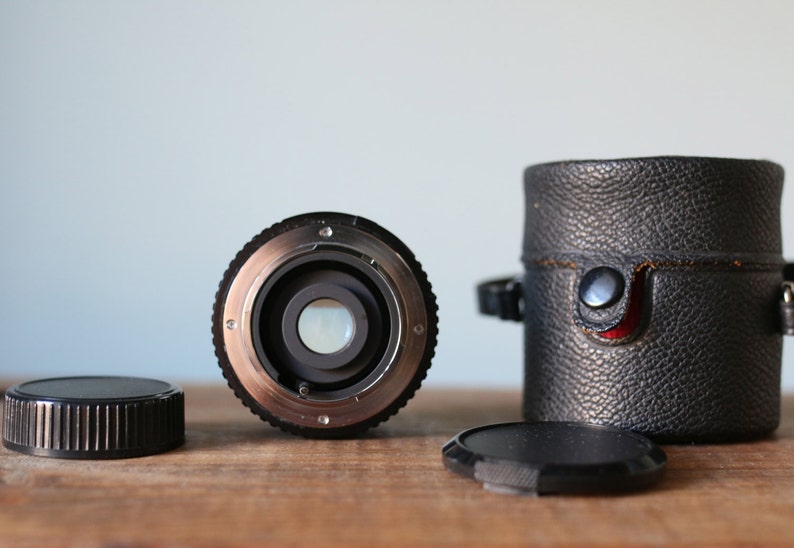May include: A black camera lens with a silver ring around the front. The lens is sitting on a wooden surface with a black leather case and a black lens cap.
