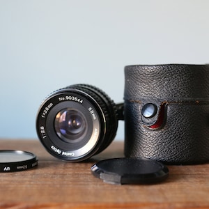 May include: A black camera lens with a 52mm filter size and a lens cap. The lens has the text "Coated Optics 1:2.8 f=28mm No. 903544 52mm" printed on it.
