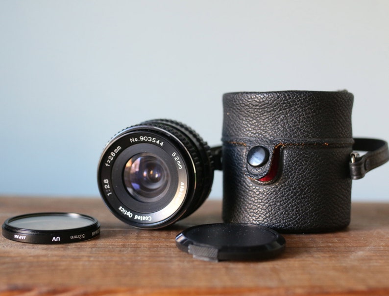 May include: A black camera lens with a 52mm filter size and a lens cap. The lens has the text "Coated Optics 1:2.8 f=28mm No. 903544 52mm" printed on it.