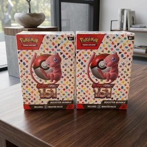 Könnte beinhalten: Zwei "Pokémon Trading Card Game" Booster-Bundles. Jede Box zeigt einen rosa Mew-Charakter mit einem Pokéball, das Logo "Scarlet & Violet 151" und enthält 6 Booster-Packs. Die Boxen stehen auf einer Holzoberfläche.