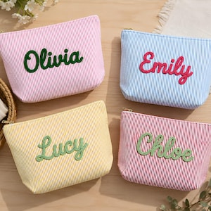 Peut inclure: Quatre trousses rectangulaires zippées en tissu rayé rose, bleu, jaune et rouge. Chaque trousse présente un prénom brodé en écriture cursive : Olivia en vert foncé, Emily en rose vif, Lucy en vert clair et Chloe en vert clair.