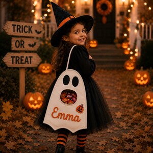 May include: A white felt ghost-shaped trick-or-treat bag featuring a cutout window filled with sweets, the name Emma embroidered in orange, and a small pumpkin patch. The bag has a long strap and is worn over a black dress with orange and black striped socks.