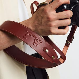May include: A dark brown leather camera strap featuring a debossed monogram logo with the letters A and J. The strap is constructed with smooth leather, secured by metallic rivets, and includes a brass-toned swivel clasp for camera attachment.