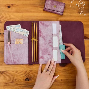 May include: A deep purple leather journal cover with a marbled texture, featuring yellow elastic bands, multiple interior pockets for cards and papers, and a matching small wallet. The journal is open, displaying slots for cards and a pen holder.