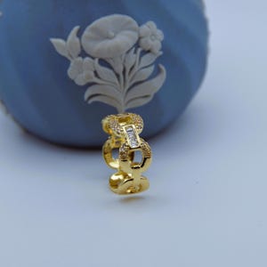 Könnte beinhalten: Ein goldfarbener Ring mit einem Kettenglied-Design. Der Ring hat einen rechteckigen, klaren Stein in der Mitte des Kettenglieds, umgeben von kleineren, klaren Steinen. Der Ring ist vor einem blauen Hintergrund platziert.