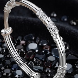 Pode incluir: Uma pulseira rígida de tom prateado com um design ajustável sobreposto e padrões florais e de folhas em relevo na superfície externa. O metal tem um acabamento polido e reflexivo e inclui uma pequena marca de contraste na lateral.