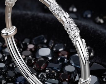 Może przedstawiać: Srebrzysta bransoletka typu bangle o regulowanej, nakładającej się konstrukcji z wytłoczonymi wzorami kwiatów i liści na zewnętrznej powierzchni. Metal ma polerowane, odblaskowe wykończenie i zawiera mały znak probierczy z boku.