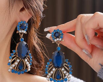 May include: A pair of ornate blue and gold earrings. Each earring features a large, oval, dark blue gemstone, surrounded by smaller blue stones and white pearls. The earrings have a vintage, chandelier style.