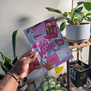 May include: A greeting card with a light blue and pink gradient background. The card features the words "Big Mama" in large pink letters, along with various stickers and graphics. The card is held in a person's hand.