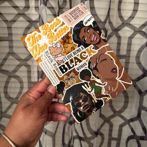 May include: A decorative card with the text "The Way Book Was Better" and "I Love Being a Black Woman". The card features illustrations of Black women, butterflies, and a Hello Kitty character. The card is held by a person.