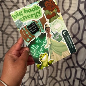 Pode incluir: Um caderno coberto de adesivos com as palavras "big book energy" e "Hello Gorgeous". Os adesivos apresentam imagens de desenhos animados de uma princesa, um sapo e outros gráficos relacionados à leitura. O caderno é segurado em uma mão.