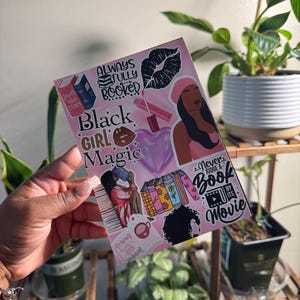 Könnte beinhalten: Ein rosa-schwarzer Kunstdruck mit den Worten "Black Girl Magic" und anderen Texten und Illustrationen. Der Druck zeigt Bilder von Büchern, Lippen und einer Frau mit rosa Kopfbedeckung. Der Druck wird von einer Person gehalten.