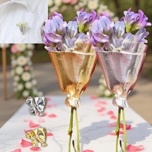 Puede incluir: Dos jarrones decorativos de cristal, uno dorado y otro plateado, cada uno con flores moradas. Los jarrones tienen forma de tulipanes estilizados. También se muestran pequeños broches plateados y dorados con forma de tulipán, junto con un pequeño prendedor floral prendido a una camisa blanca.