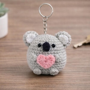May include: A gray crocheted koala keychain with a pink heart on its chest. The koala has black eyes and a black nose. The keychain is attached to a silver keyring. A succulent plant in a white pot and a set of keys are in the background.