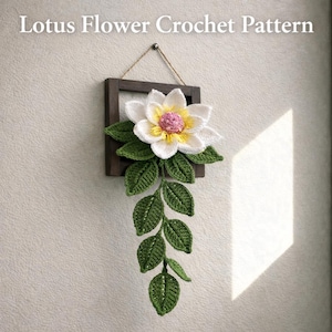 Op de afbeelding: Een gehaakte lotusbloem wanddecoratie met een witte bloem, gele kern en roze accenten, omlijst in een donkerhouten vierkant. Groene bladeren hangen naar beneden. De tekst "Lotus Flower Crochet Pattern" staat bovenaan.