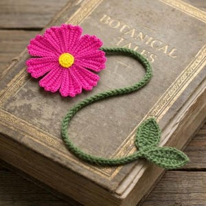 Può includere: Segnalibro all'uncinetto fatto a mano con un vivace fiore rosa e un centro giallo. Il fiore è collegato a uno stelo e foglie verdi. Il segnalibro è appoggiato su un libro antico con il titolo "Botanical Tales" in lettere dorate.