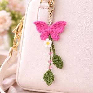 May include: A pink crochet butterfly keyring with a white flower, green leaves, and pink beads. The butterfly is attached to a gold-coloured clasp, hanging from a white handbag. The background is blurred.