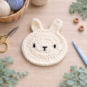 May include: A handmade, off-white crochet bunny coaster with black eyes and a black 'T' shaped nose. The coaster is round with bunny ears. Other craft supplies, including scissors, yarn, and a crochet needle, are in the background.