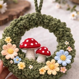 Op de afbeelding: Een handgemaakte gehaakte krans met een groene basis, versierd met kleurrijke bloemen en twee rood-witte paddenstoel figuren. De krans heeft een lus om op te hangen, een charmant decoratief stuk.