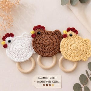 May include: Three handmade crochet chicken towel holders with wooden rings. The chickens are white, brown, and light yellow, each featuring a red comb, yellow beak, and black eye. A rectangular tag reads: HANDMADE CROCHET - CHICKEN TOWEL HOLDERS.