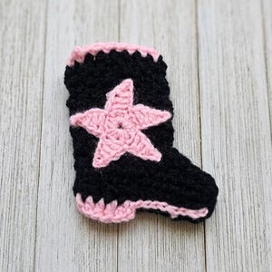 May include: A black crochet boot with a pink star and trim. The handmade boot has a star in the center and pink trim around the top and sole. The boot is set against a light wood background.