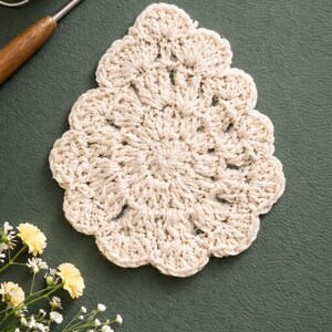 May include: A cream-colored, handmade crochet coaster with a scalloped edge. The coaster is circular with a floral design in the center. A ball of yarn and a crochet hook are in the background.