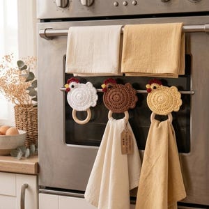 May include: Three crochet chicken towel holders in white, brown, and yellow, each featuring a wooden ring. Two plain rectangular kitchen towels hang from the oven handle, while two additional towels are suspended from the chicken-shaped holders.
